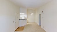 This single-floor residence offers an open and airy layout that immediately draws the eye upward to its impressive vaulted ceiling, which lends a generous sense of height and spaciousness throughout. A ceiling fan centered in the main living area provides both function and a finishing touch to the room's clean, neutral aesthetic.

The living space flows naturally toward an open-concept kitchen, which is defined by a raised breakfast bar-style counter with a dark countertop that provides an ideal spot for casual dining or meal prep. The kitchen is outfitted with white cabinetry, white appliances including a refrigerator and range, and benefits from a skylight above that brings in additional natural light, keeping the space bright and welcoming.

The flooring transitions from hardwood-style planks near the entryway to plush, light-colored carpet that covers the main living area, adding warmth and comfort underfoot. The walls are finished in a soft, neutral white tone throughout, creating a clean backdrop that suits a variety of décor styles.

A doorway off the living area leads further into the residence, suggesting additional private rooms beyond. The front entry features a solid white door, providing a clear and practical threshold between the interior and the outside world. Overall, the space is characterized by its brightness, open sightlines, and versatile, move-in-ready layout.