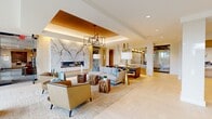 This sophisticated single-floor amenity space offers an impressive and thoughtfully designed environment, encompassing a generous footprint that accommodates a living room, dining area, and a fully equipped kitchen. The entry foyer sets an elegant tone with light wood herringbone flooring, glass architectural accents, and clean modern lines that welcome visitors into the space.

The central living area is an architectural showpiece, anchored by a dramatic floor-to-ceiling marble fireplace surround that commands immediate attention. A coffered ceiling with a warm gold inlay adds depth and grandeur, while a contemporary geometric chandelier provides focused ambient lighting. Plush neutral seating, accented by organic wood side tables, creates a refined yet inviting lounge atmosphere.

The kitchen is outfitted with premium stainless steel appliances, light wood cabinetry, open display shelving, and crisp white countertops. Floor-to-ceiling windows in this area flood the space with natural light and offer views of the lush landscaped surroundings outside.

Additional seating alcoves feature soft textured wall panels with warm accent lighting, offering intimate gathering spots throughout. Throughout the space, light hardwood flooring, recessed lighting, and a neutral palette of warm whites, golds, and natural tones create a cohesive, upscale ambiance ideal for communal use.