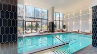 Located at 1180 Rue de Bleury in the heart of Montréal, this sophisticated indoor pool and amenity space offers a resort-caliber experience within an urban setting. The generously sized pool area is defined by soaring double-height ceilings and expansive floor-to-ceiling glazing that floods the space with natural light and frames striking views of the surrounding downtown skyline. The pool itself is bordered by refined stone coping and elegant tile surrounds, complemented by a poolside deck furnished with wood-framed lounge chairs and marble accent tables for a refined, contemporary aesthetic. Dramatic dark hexagonal tile columns serve as bold architectural focal points, contrasting beautifully with the warm natural wood ceiling panels and crisp white walls adorned with softly backlit ribbed paneling. A glass-partitioned fitness area is seamlessly integrated into the layout, maintaining visual openness throughout. Sliding glass doors connect the interior to a landscaped outdoor terrace, where wicker daybed loungers are set amid lush greenery and planters against a backdrop of Montréal's iconic cityscape. The overall ambiance balances warmth and sophistication, delivering a tranquil retreat designed for relaxation and wellness in the heart of the city.