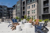 Courtyard 2 is a thoughtfully designed outdoor communal space located in Meridian, Idaho, enclosed by contemporary multi-story buildings featuring a modern architectural palette of brick, horizontal siding, and dark accent panels. The courtyard offers a generous open-air environment bathed in abundant natural sunlight, anchored by a striking wood-and-lattice pergola at its center, strung with overhead bistro lights that lend a welcoming ambiance both day and evening.

The space is organized into distinct activity zones. A wide concrete plaza serves as the main gathering area, furnished with an assortment of outdoor seating arrangements including white lounge sofas with yellow cushions, Adirondack chairs, and high-top dining tables with chairs and umbrellas. A bocce ball court adds a recreational element to the space. Raised concrete planters with young trees and ornamental landscaping provide natural accents throughout.

Surrounding the hardscaped areas, manicured lawn sections offer open green space, complemented by well-maintained landscaping with shrubs and perennials. A paved walkway flanked by greenery connects the courtyard to adjacent areas of the property, ensuring easy pedestrian flow. The enclosing buildings feature private balconies overlooking the courtyard, creating an integrated indoor-outdoor community experience.
