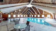 This single-story indoor pool facility is defined by its striking architectural design, centered around a soaring vaulted ceiling clad in warm wood planking and supported by bold white structural trusses. The peaked roofline creates an impressive sense of height and openness throughout the space. A series of pendant globe lights hang at varying levels along the trusses, providing ambient illumination that complements the natural light streaming in through the numerous windows lining the perimeter walls. These windows also offer views of the surrounding outdoor greenery, enhancing the connection to the natural environment.

The centerpiece of the facility is the generously sized rectangular swimming pool, with clear blue water visible beneath the well-lit interior. Surrounding the pool is an expansive deck finished in large-format stone tile laid in an irregular flagstone pattern, lending a rustic yet refined aesthetic to the space. The deck area is furnished with round glass-top tables and sling-back chairs, providing comfortable spots for relaxation before or after a swim. The warm wood tones of the ceiling, combined with the stone flooring and abundant natural light, create an inviting and resort-like ambiance throughout.