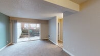 This single-floor residence is thoughtfully laid out across several well-defined areas. The main living space is anchored by large sliding glass doors that provide direct access to an outdoor patio area, allowing natural light to fill the room and offering views of the exterior surroundings. The living area features wall-to-wall carpet and a textured ceiling, with a structural beam detail adding architectural character. Flooring transitions from carpet to hard surface wood-tone planks near the entry and kitchen zones.

The kitchen is equipped with warm-toned wooden cabinetry, granite-patterned countertops, a stainless steel single-basin sink, and a window with blinds that brings in additional natural light. A tile floor and hardwood-style flooring complement the functional layout.

The bedrooms are carpeted and feature neutral-toned walls, with one room showcasing a soft sage-green accent wall and a sliding door for outdoor access. A second bedroom offers a window with blinds and a wall-mounted heating unit.

The bathroom includes a full tub-shower combo with a molded surround featuring built-in corner shelving, paired with clean white fixtures throughout. The overall palette remains neutral and understated, creating a calm, functional living environment.
