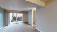 This single-floor residence is thoughtfully laid out across several well-defined areas. The main living space is anchored by large sliding glass doors that provide direct access to an outdoor patio area, allowing natural light to fill the room and offering views of the exterior surroundings. The living area features wall-to-wall carpet and a textured ceiling, with a structural beam detail adding architectural character. Flooring transitions from carpet to hard surface wood-tone planks near the entry and kitchen zones.

The kitchen is equipped with warm-toned wooden cabinetry, granite-patterned countertops, a stainless steel single-basin sink, and a window with blinds that brings in additional natural light. A tile floor and hardwood-style flooring complement the functional layout.

The bedrooms are carpeted and feature neutral-toned walls, with one room showcasing a soft sage-green accent wall and a sliding door for outdoor access. A second bedroom offers a window with blinds and a wall-mounted heating unit.

The bathroom includes a full tub-shower combo with a molded surround featuring built-in corner shelving, paired with clean white fixtures throughout. The overall palette remains neutral and understated, creating a calm, functional living environment.