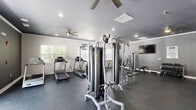 Located at Chanticleer Village in Myrtle Beach, South Carolina, this well-equipped fitness center offers a comprehensive workout environment within a single, open-plan space. The room features durable rubber flooring throughout, providing both safety and comfort during exercise sessions. Ceiling fans and recessed lighting ensure the space remains well-lit and comfortable during workouts.

The cardio zone is equipped with multiple treadmills and elliptical trainers, positioned along windowed walls that allow ample natural light to fill the room. A multi-station cable machine serves as the centerpiece of the strength training area, complemented by additional weight machines and a dedicated free weights section with a tiered dumbbell rack. A weight bench and seated exercise equipment round out the strength training offerings.

A separate stretch and flex area features full-length mirrored walls that visually expand the space, along with padded seating benches and yoga mat storage for warm-up and cool-down routines. A wall-mounted flat-screen television provides entertainment during workouts. Elegant French doors with an arched transom window offer a bright, welcoming entry point, while additional windows along the perimeter bring in natural light and views of the surrounding community. The overall design is clean, functional, and modern, creating an inviting atmosphere for residents pursuing an active lifestyle.