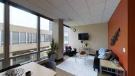 This single-floor space offers a functional and comfortable layout with a strong urban character. The living area is a standout feature, anchored by large floor-to-ceiling windows along two walls that flood the interior with natural light while framing views of the surrounding cityscape. The drop-tile ceiling adds a clean, structured finish overhead.

The living area is thoughtfully arranged with a plush sofa and a leather armchair, both positioned to face a wall-mounted flat-screen television. A light-toned area rug grounds the seating arrangement, complementing the neutral beige walls and creating a warm, inviting atmosphere. A compact side table between the seating pieces provides a practical surface, while decorative greenery — including a tall floor plant — adds life and color to the space.

A dining or work area is visible toward the front of the space, featuring a light wood table and chairs, making it suitable for both meals and work-from-home activities. The overall design balances practicality with comfort, and the abundance of natural light combined with city views gives the space a dynamic, connected-to-the-urban-environment feel.