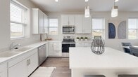 This thoughtfully designed three-story home offers a clean, contemporary aesthetic with carefully selected finishes throughout. The entry foyer welcomes residents with neutral-toned carpet, crisp white walls, and a paneled front door with a transom window that invites natural light into the space.

The main living level features an open-concept layout connecting the living room and kitchen. The living room is bright and airy, with multiple windows providing abundant natural light and views of the surrounding landscape, complemented by recessed lighting and warm hardwood-style flooring. The kitchen is a standout, boasting white shaker cabinetry, quartz countertops, a central island with bar seating, stainless steel appliances, and pendant lighting overhead.

The bedrooms are comfortably appointed with plush carpeting, ceiling fans, and windows that bring in natural light. The bathrooms are finished with tiled flooring, white vanity cabinetry, quartz countertops, and large mirrors. One bathroom features a dedicated vanity area with built-in storage pull-outs, while another includes a walk-in shower with a classic curtain rod.

Connecting all three floors, the staircase is fitted with patterned, multi-tone carpeting and white-painted risers with dual handrails, offering both safety and visual appeal. The overall ambiance is modern, clean, and welcoming throughout.
