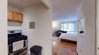 This single-floor studio is thoughtfully laid out to maximize every inch of available space. The open main area seamlessly combines sleeping and living functions, centered around warm parquet wood flooring that runs throughout. A large window dressed with vertical blinds allows generous natural light to fill the room, creating a bright and airy atmosphere. The living area accommodates a bed, a sofa, a coffee table, and a dining set with chairs, while a wooden dresser provides additional storage. A compact kitchen is positioned just off the main living area, featuring warm oak cabinetry that extends both above and below the counter, a stainless steel gas range, a double sink, and stone-look countertops — offering ample prep and storage space. A small pass-through window connects the kitchen area to the main living space, adding an open feel. The bathroom includes a vanity with matching stone-look countertop and warm wood cabinetry, complemented by a dedicated closet area with shelving and hooks for organized storage. The unit's clean white walls and consistent flooring throughout create a cohesive, neutral palette that lends itself well to a variety of interior styles.
