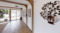 This single-floor lobby space presents a warm and inviting atmosphere rooted in a Southwestern architectural style. The entryway is defined by ornate arched doorways trimmed in a rich tan tone, lending a distinctive character to the interior. Decorative elements such as a wrought-iron leaf wall sculpture and framed artwork add thoughtful touches throughout.

The flooring transitions between terracotta-style ceramic tile near the entrance corridor and textured carpet in the lounge area, clearly delineating zones within the space. Large floor-to-ceiling glass panels and sliding glass doors along the perimeter flood the interior with natural light while offering open views of mature trees, lush greenery, and the surrounding outdoor landscape.

A casual seating area features a round dark table with matching chairs, providing a comfortable spot for guests to gather. A Coca-Cola vending machine is conveniently positioned nearby. Exposed wood beam accents along the ceiling add warmth and visual interest, complementing the overall earthy color palette of white walls and brown trim.

The front entrance is equipped with a full glass door and automatic door mechanism, opening onto a brick-paved exterior walkway flanked by landscaping. The corridor leading inward is bright and airy, with additional framed art and a decorative half-door gate contributing to the welcoming character of the space.