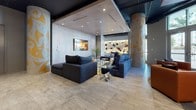 This dynamic single-floor amenity lounge is designed to impress, blending urban artistry with sophisticated interior design. The space is anchored by a striking floor-to-ceiling graffiti-style mural bursting with vivid color and expressive imagery, serving as an unmistakable focal point near the entrance. Floor-to-ceiling windows along the perimeter flood the interior with natural light, complemented by sheer drapery that softens the atmosphere while maintaining an open connection to the outdoors.

The lounge area features plush navy sectional sofas arranged around glass-topped coffee tables atop a richly patterned area rug, creating an inviting setting for gathering and relaxation. Decorative columns adorned with intricate golden motifs add architectural character throughout. A cozy seating vignette with warm cognac leather chairs and a low round table provides an intimate contrast near the entrance.

A dedicated bar-height workspace station is equipped with sleek black counter seating, a mounted flat-screen display, and sculptural orbital pendant lighting overhead — ideal for casual meetings or focused work. A fireplace with an art photograph above it anchors a quieter lounge alcove, while a curated display shelf and additional seating complete the space. The concrete ceiling with recessed lighting and exposed structural elements lend an industrial-chic character throughout.