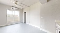 This single-floor unit offers a thoughtfully organized layout across three rooms, making efficient use of its compact footprint. The main living area is bathed in natural light from a large sliding window and is equipped with a wall-mounted mini-split air conditioning unit and a ceiling fan, ensuring year-round comfort. The painted concrete floors throughout lend the space a clean, modern aesthetic that ties each room together cohesively.

The kitchen occupies a dedicated nook and features taupe shaker-style cabinetry, a stainless steel refrigerator, a freestanding gas range with an overhead range hood, and a single-basin sink — providing all essential cooking amenities in a streamlined layout.

The bedroom offers a quiet retreat with its own ceiling fan and a window framing views of palm trees and the surrounding urban skyline, filling the room with soft, diffused light. A utility closet accessible from this room houses the water heater and shelving for additional storage.

The bathroom is finished with white subway tile surrounding a full soaking tub and shower, complemented by wood-look tile flooring, a pedestal sink, and brushed gold fixtures that add a warm, refined touch. The textured white walls throughout the unit contribute to a bright, airy ambiance that maximizes the sense of space.