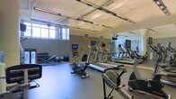This single-floor fitness center offers a practical and well-organized workout environment designed to accommodate a range of exercise needs. The space is bright and open, benefiting from natural light that filters in through windows along one wall, complemented by rows of overhead fluorescent lighting mounted to a white-painted ceiling with exposed structural beams.

The cardio section is a central feature, housing multiple treadmills equipped with individual screens, alongside stationary and recumbent bikes. A full-length mirror runs along one wall, visually expanding the space and allowing users to monitor their form during workouts. The mirror also reflects the mounted flat-screen televisions, adding to the functionality of the cardio area.

Along the opposite wall, a dedicated free weights section includes a rack of dumbbells, a weight bench, and stability balls, providing a versatile strength training area. The flooring throughout is a durable, cushioned rubber surface suited for heavy equipment and high-impact activity. The overall ambiance is clean, functional, and well-maintained, making it a capable facility for both cardio and strength training routines.