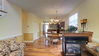 This two-story home presents a welcoming main level defined by rich hardwood flooring that flows seamlessly throughout the open-concept living and dining areas. The dining space is anchored by a round wooden table with cushioned chairs, positioned beneath an elegant chandelier that adds a refined focal point to the room. Natural light streams in through multiple windows fitted with blinds, creating a bright and airy atmosphere complemented by warm yellow-toned walls with classic white wainscoting detail.

A glass-enclosed fireplace serves as a stylish design element, providing both visual warmth and a practical heating feature at the edge of the living area. The kitchen, visible from the dining space, features dark espresso cabinetry, stainless steel appliances including a refrigerator and over-the-range microwave, and a functional layout suited for everyday cooking. A peninsula counter connects the kitchen to the dining area, enhancing the open flow of the space.

The layout includes a hallway leading to additional rooms, suggesting a well-organized floor plan. Overall, the main level balances casual comfort with tasteful design details throughout.