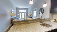 This single-floor residence in Windsor offers a thoughtfully designed open-concept layout that seamlessly connects the kitchen, dining, and living areas. The kitchen is a standout feature, showcasing expansive granite countertops with a speckled finish, a stainless steel sink with a modern arc faucet, and stylish pendant lighting that adds a warm, contemporary touch overhead. The kitchen flows naturally into the dining and living spaces, creating an inviting atmosphere ideal for both everyday living and entertaining. The living area is bathed in natural light, courtesy of large floor-to-ceiling windows and elegant French doors that frame outdoor views and allow sunlight to fill the room throughout the day. Cool, soft blue-gray walls lend a calm and cohesive tone throughout the space, complementing the light-toned flooring and neutral furnishings. The open sightlines from the kitchen through to the living area enhance the sense of spaciousness, making the layout feel airy and connected. Pendant and floor lamps provide layered lighting options for the evening hours. Overall, this residence combines modern finishes with a relaxed, welcoming ambiance that is both functional and visually appealing.