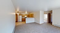 This single-floor space is designed with an open-concept layout that seamlessly connects the kitchen and living area. The kitchen is equipped with honey-toned wood cabinetry offering ample storage, a white refrigerator, a range, and a peninsula that serves as a natural divider between the cooking and living spaces. The flooring in the kitchen consists of neutral tile, providing a practical and easy-to-maintain surface. A ceiling fan with a light fixture is centrally positioned over the kitchen area, contributing to both ventilation and ambient lighting.

The living area transitions smoothly from the kitchen, featuring wall-to-wall carpeting in a neutral gray tone that adds warmth underfoot. A window along the far wall allows natural light to fill the space. The walls throughout are painted in a clean, light neutral tone, enhancing the sense of openness. A baseboard heater runs along one wall, indicating a functional heating system integrated into the unit. The overall layout is straightforward and practical, offering a comfortable, low-maintenance living environment suited to everyday use.