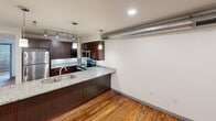 This two-floor apartment at 1101 Hawthorne Lane in Charlotte's 28205 neighborhood showcases an industrial-chic aesthetic throughout. The main level features rich, reclaimed hardwood floors, striking exposed brick accent walls, and an open-concept kitchen outfitted with dark wood cabinetry, granite countertops, stainless steel appliances, a double-basin sink, and pendant lighting. Exposed ductwork and a structural column reinforce the loft-style character. The upper level offers lighter hardwood flooring, wood-plank ceilings with exposed beams, and a window providing natural light. The bathroom includes a granite-topped double vanity, a full tub/shower combo, and accessible grab bars.