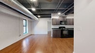 Located at 248 N Santa Fe Ave in Salina, Kansas, this single-floor urban loft offers a distinctive industrial-chic aesthetic that blends raw architectural character with modern comfort. Warm hardwood flooring runs throughout the space, complementing the dramatic exposed ceiling with dark-painted beams, metal ductwork, and recessed lighting that together create an open, loft-style ambiance.

The combined dining, kitchen, and living area forms the heart of the unit, bathed in natural light from multiple large windows framed against whitewashed brick walls. The well-appointed kitchen features dark wood cabinetry, dark countertops, a full suite of black appliances including a refrigerator, electric range, and microwave, all set beneath the striking open ceiling.

The two bedrooms continue the loft's industrial character, each showcasing the same exposed beam ceilings, ceiling fans, arched or rectangular windows with views to the surrounding streetscape, and rich hardwood floors. The primary bedroom offers generous proportions with ample room to furnish comfortably. Storage is notably well-served with four closets throughout, including a spacious walk-in closet with wire shelving systems.

The bathroom features tile flooring and a clean, functional layout. A dedicated utility room and a connecting hallway round out this thoughtfully designed urban residence.