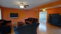 This single-floor space centers around a lively and welcoming living room distinguished by its bold, warm orange walls that create an energetic and inviting atmosphere. The room is furnished with dark wicker-style seating, including a sofa adorned with earth-toned accent pillows and a matching armchair, arranged around a rectangular wooden coffee table. A wall-mounted flat-screen television anchors one side of the room, making it a practical and functional entertainment area. Overhead, a ceiling fan with integrated lighting provides both air circulation and ambient illumination, contributing to the room's comfortable feel. The flooring throughout is smooth hardwood, adding a clean and polished look to the space. A partial column detail subtly divides the living area, adding a touch of architectural interest. At the far end of the room, floor-to-ceiling vertical blinds frame a sliding glass door that opens to an outdoor area, allowing natural light to filter into the space and offering a visual connection to the exterior. A second ceiling fan is visible beyond the dividing column, suggesting a seamlessly flowing, open layout throughout the main living area.