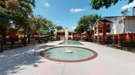 Located at 4720 Reading Rd in Rosenberg, Texas, this residential community showcases a classic architectural style defined by rich red brick exteriors, arched entryways, and traditional chimney details. The buildings blend brick and siding construction, giving the community a warm, established character that is further enhanced by mature trees and colorful crepe myrtle plantings throughout the grounds.

The outdoor amenity area serves as a central gathering point, featuring a well-maintained swimming pool alongside a charming circular wading fountain with a center jet — a standout detail that adds visual interest and a sense of leisure to the space. The pool area is enclosed by ornamental iron fencing, providing both safety and a refined aesthetic. A shaded canopy structure nearby offers a comfortable retreat from the Texas sun.

The landscaped grounds are thoughtfully maintained, with green lawns, mature shade trees, and flowering shrubs framing the walkways and building exteriors. Decorative iron fencing with circular detailing defines pathways and common areas, contributing to the community's cohesive design. The single-story layout of the featured unit offers easy accessibility, with a brick lower exterior and light siding above. Overall, the community presents a well-established, inviting environment with practical amenities and attractive curb appeal in a suburban Rosenberg setting.