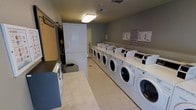 This shared laundry facility is a practical, single-floor space designed to accommodate the laundry needs of multiple residents efficiently. The room is laid out with a long row of commercial-grade front-loading washing machines lining one wall, paired with a corresponding row of dryers stacked or positioned nearby, offering generous capacity for simultaneous use. Overhead fluorescent lighting provides bright, even illumination throughout the space, ensuring a well-lit and functional environment. The walls are painted in a neutral gray tone, lending the room a clean and utilitarian appearance. Light-colored tile flooring runs throughout, contributing to easy maintenance and a tidy overall look. A dedicated folding counter provides a flat, spacious surface for sorting and folding laundry. A utility cabinet offers additional storage, while posted instructional signage on the walls ensures users can operate the machines correctly. The room also features a coin-operated or card-based payment station, facilitating convenient self-service use. Ventilation is provided through ceiling vents, supporting a comfortable atmosphere during operation. Overall, this laundry room is thoughtfully organized to maximize efficiency and ease of use for its residents.