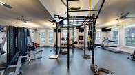 Located at 4531 Langdon Dr in Durham, NC, this single-floor fitness facility is thoughtfully designed to accommodate a wide range of workout styles and training goals. The space is divided into distinct functional zones, connected by a hallway, and is finished throughout with durable rubber and hardwood-style flooring suited for high-performance use.

The main gym area is expansive and airy, featuring soaring ceilings, ceiling fans, and an abundance of natural light streaming through multiple white-framed windows. Wall-mounted TVs provide entertainment or instructional content throughout. This zone is outfitted with a large multi-functional power rack system by Body-Solid, complete with TRX suspension trainers, a heavy punching bag, and battle ropes. Additional equipment includes a full dumbbell rack, cable machines, weight benches, treadmills, a rowing machine, an AbSolo trainer, and a stationary bike. Full-length mirrors line portions of the walls, enhancing the sense of space and allowing for form monitoring.

A separate studio area features warm hardwood-style flooring, soft sage-green walls, ample window light, and a ceiling fan. This room houses a row of spin bikes alongside stability balls and step platforms, creating an ideal setting for group cycling or low-impact training sessions. The overall atmosphere is clean, modern, and energizing.