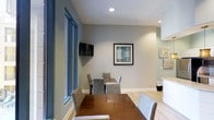 The game room at 1670 Kettner in San Diego is a thoughtfully designed communal space that balances functionality with a clean, modern aesthetic. The room features light gray walls accented by white baseboards, creating a neutral and inviting atmosphere. Recessed ceiling lighting provides consistent, even illumination throughout the space. A large wooden dining or game table serves as the centerpiece, surrounded by upholstered gray chairs that offer comfortable seating for multiple guests. A wall-mounted flat-screen television is positioned for easy viewing from the table, making the space well-suited for both casual gaming and social gatherings. Framed artwork adorns the walls, adding a decorative touch that complements the contemporary design. Light hardwood-style flooring runs throughout, contributing to the room's bright and open feel. Floor-to-ceiling windows along one side allow natural light to filter in, enhancing the airy ambiance. An adjacent area, partially visible, suggests additional amenity spaces nearby. Overall, the game room offers a versatile and well-appointed environment designed for leisure and community engagement.