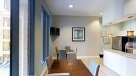 The game room at 1670 Kettner in San Diego is a thoughtfully designed communal space that balances functionality with a clean, modern aesthetic. The room features light gray walls accented by white baseboards, creating a neutral and inviting atmosphere. Recessed ceiling lighting provides consistent, even illumination throughout the space. A large wooden dining or game table serves as the centerpiece, surrounded by upholstered gray chairs that offer comfortable seating for multiple guests. A wall-mounted flat-screen television is positioned for easy viewing from the table, making the space well-suited for both casual gaming and social gatherings. Framed artwork adorns the walls, adding a decorative touch that complements the contemporary design. Light hardwood-style flooring runs throughout, contributing to the room's bright and open feel. Floor-to-ceiling windows along one side allow natural light to filter in, enhancing the airy ambiance. An adjacent area, partially visible, suggests additional amenity spaces nearby. Overall, the game room offers a versatile and well-appointed environment designed for leisure and community engagement.