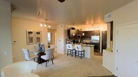 This well-appointed single-floor residence offers an open-concept layout that seamlessly connects the living, dining, and kitchen areas into one cohesive and inviting space. The modern kitchen is a standout feature, outfitted with dark espresso cabinetry, a striking mosaic tile backsplash, stainless steel appliances including a refrigerator and microwave, and a peninsula breakfast bar with seating for multiple guests. Pendant lighting and track lighting illuminate the kitchen, adding both functionality and a warm ambiance. The dining area comfortably accommodates a glass-top table surrounded by elegant upholstered chairs, set beneath a decorative multi-bulb light fixture that enhances the contemporary aesthetic. The adjacent living area is carpeted in plush neutral tones, providing a soft, comfortable foundation that contrasts nicely with the hard flooring in the kitchen and entry zones. Warm, ambient lighting throughout the space creates an inviting atmosphere suited for both everyday living and entertaining. A hallway leads to the private areas of the residence, and a window allows natural light to filter in, brightening the overall interior. The cohesive design palette of dark wood tones, neutral walls, and modern fixtures gives the space a polished and sophisticated character.