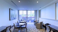 This contemporary single-floor space is designed as a versatile communal lounge, blending functionality with a clean, modern aesthetic. The interior is defined by crisp white walls and recessed lighting that casts a bright, even illumination throughout the room, contributing to an open and welcoming atmosphere.

The space is thoughtfully arranged with two distinct seating zones. A dining or work area along one side features dark rectangular tables paired with upholstered side chairs, offering an ideal setting for casual meetings or individual work. On the opposite side, a relaxed lounge area is furnished with patterned accent chairs and small side tables, providing a comfortable space for informal conversation or quiet reading.

A patterned carpet in muted tones of gray and blue anchors the floor, adding visual interest while complementing the overall neutral color palette. A large piece of abstract wall art brings a touch of personality to the space.

One of the most notable features is the floor-to-ceiling glass wall and sliding doors that flood the interior with natural light and offer a seamless connection to an outdoor patio. The patio, visible through the expansive glazing, is landscaped with lush greenery and additional seating, making it an attractive extension of the indoor environment. The overall ambiance is calm, professional, and inviting.