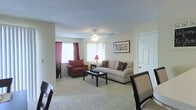 This single-floor residence offers a comfortable and functional layout designed for everyday living. The living room serves as the heart of the space, featuring plush wall-to-wall carpeting that adds warmth underfoot. Multiple windows dressed with rich burgundy curtains allow natural light to filter in, creating a bright and inviting atmosphere. A ceiling fan with an integrated light fixture is centrally positioned, providing both airflow and ambient lighting throughout the room. The living area comfortably accommodates a full seating arrangement, with ample space for a sofa, armchair, and a coffee table, making it well-suited for both relaxing and entertaining. Neutral, light-toned walls provide a versatile backdrop that complements a variety of décor styles. A framed piece of wall art adds a touch of character to the space. The textured ceiling contributes a classic residential feel. A nearby door hints at additional rooms or entry points, suggesting a practical and connected floor plan. Overall, the space presents a cozy, well-lit, and neatly arranged single-level home with a warm ambiance and a functional, traditional design sensibility.