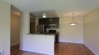 This single-floor space showcases a thoughtfully designed open-concept layout that seamlessly connects the kitchen and living area. The kitchen is equipped with stainless steel appliances, including a refrigerator, microwave, and range, complemented by rich dark cabinetry that extends to the upper walls for generous storage. A peninsula with granite countertops serves as both a functional workspace and a natural divider between the kitchen and the adjoining living area, offering casual seating potential. A stylish pendant light hangs above the living space, adding a decorative focal point and ambient illumination, while a flush-mount ceiling fixture lights the kitchen zone. The flooring throughout features warm hardwood-style planks that enhance the cohesive, modern aesthetic. The textured ceiling adds depth overhead. The open flow between spaces creates a practical and welcoming environment, making the layout well-suited for everyday living and entertaining. A hallway visible in the background suggests additional rooms beyond the main living area.