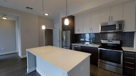 This single-floor space showcases a thoughtfully designed, modern interior centered around a striking contemporary kitchen. The kitchen is a standout feature, boasting a generous island with a pristine light quartz countertop that serves as both a functional workspace and a social hub. Elegant pendant lights hang overhead, casting a warm glow that complements the room's clean aesthetic.

The cabinetry combines crisp white upper cabinets with rich dark espresso lower cabinets, creating a bold two-tone contrast that adds depth and sophistication. A dark subway tile backsplash ties the design together, adding texture and a refined visual element. Stainless steel appliances — including a French door refrigerator with an ice and water dispenser, a gas range, a dishwasher, and an over-the-range microwave — provide a high-end, cohesive look throughout.

The space flows naturally from the kitchen into adjoining areas, offering an open and airy feel. Dark hardwood-style flooring grounds the interior, providing a warm counterpoint to the lighter cabinetry and countertops. A hallway visible from the kitchen leads to additional areas of the space, suggesting a practical and well-organized layout. Overall, the design balances modern aesthetics with everyday functionality.