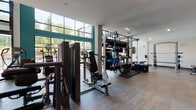 This two-story fitness center at 500 Buford Hwy is a well-appointed, modern facility designed to accommodate a full range of workout styles. The main floor is anchored by soaring double-height windows framed in dark steel, flooding the space with natural light and offering direct views of the outdoor pool and surrounding greenery. A large garage-style window wall can open to connect the interior with the outdoor environment, enhancing the airy, open atmosphere. The ground level houses an extensive selection of cardio equipment, including rowing machines, ellipticals, stair climbers, and spin bikes, all positioned to take advantage of the outdoor views. A dedicated free weights area features a full rack of dumbbells, stability balls, and mirrored walls. Matrix strength machines and a functional training rig with cables and suspension systems provide comprehensive strength training options. Warm wood-toned flooring runs throughout, complemented by crisp white walls and teal accent walls. A staircase with stylish metal railings leads to the second floor, which features additional cardio machines, Matrix treadmills, and a standout climbing wall, offering a unique and dynamic training option. Decorative plants and curated artwork add a polished, welcoming touch throughout the facility.