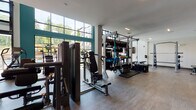 This two-story fitness center at 500 Buford Hwy is a well-appointed, modern facility designed to accommodate a full range of workout styles. The main floor is anchored by soaring double-height windows framed in dark steel, flooding the space with natural light and offering direct views of the outdoor pool and surrounding greenery. A large garage-style window wall can open to connect the interior with the outdoor environment, enhancing the airy, open atmosphere. The ground level houses an extensive selection of cardio equipment, including rowing machines, ellipticals, stair climbers, and spin bikes, all positioned to take advantage of the outdoor views. A dedicated free weights area features a full rack of dumbbells, stability balls, and mirrored walls. Matrix strength machines and a functional training rig with cables and suspension systems provide comprehensive strength training options. Warm wood-toned flooring runs throughout, complemented by crisp white walls and teal accent walls. A staircase with stylish metal railings leads to the second floor, which features additional cardio machines, Matrix treadmills, and a standout climbing wall, offering a unique and dynamic training option. Decorative plants and curated artwork add a polished, welcoming touch throughout the facility.