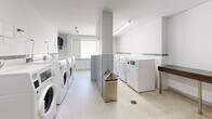 Located at 99 Dowling in Toronto's vibrant Parkdale neighbourhood, this shared laundry facility offers a clean, well-organized space designed for efficient, everyday use. The room is finished with light-toned ceramic floor tiles and crisp white walls accented by a horizontal tile border, creating a bright and hygienic environment. Overhead fluorescent lighting ensures the space is well-lit throughout.

Multiple commercial-grade front-loading washing machines and top-loading dryers are arranged along the walls, providing ample capacity for building residents. A dedicated utility sink with wood-tone cabinetry offers a convenient hand-washing or pre-treatment station. Stainless steel waste receptacles are thoughtfully placed throughout for easy disposal.

A wall-mounted television is installed near the window area, allowing residents to stay entertained during their laundry cycle. A frosted window allows natural light to filter in while maintaining privacy. Payment and operational instructions are posted near the entrance, alongside a coin-operated payment terminal for machine access. A wall-mounted fire extinguisher and storage cabinet are also present near the entrance door, reflecting attention to safety and practicality. The overall layout is functional and well-maintained, making this facility a convenient shared amenity for residents of the building.