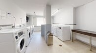 Located at 99 Dowling in Toronto's vibrant Parkdale neighbourhood, this shared laundry facility offers a clean, well-organized space designed for efficient, everyday use. The room is finished with light-toned ceramic floor tiles and crisp white walls accented by a horizontal tile border, creating a bright and hygienic environment. Overhead fluorescent lighting ensures the space is well-lit throughout.

Multiple commercial-grade front-loading washing machines and top-loading dryers are arranged along the walls, providing ample capacity for building residents. A dedicated utility sink with wood-tone cabinetry offers a convenient hand-washing or pre-treatment station. Stainless steel waste receptacles are thoughtfully placed throughout for easy disposal.

A wall-mounted television is installed near the window area, allowing residents to stay entertained during their laundry cycle. A frosted window allows natural light to filter in while maintaining privacy. Payment and operational instructions are posted near the entrance, alongside a coin-operated payment terminal for machine access. A wall-mounted fire extinguisher and storage cabinet are also present near the entrance door, reflecting attention to safety and practicality. The overall layout is functional and well-maintained, making this facility a convenient shared amenity for residents of the building.