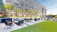 This expansive outdoor courtyard amenity space is thoughtfully designed to offer a variety of activity zones within a single cohesive environment. The courtyard is framed by a striking multi-story building facade that combines warm red brick and modern gray siding, punctuated by large steel-framed windows that allow interior spaces to connect visually with the outdoors.

The ground plane alternates between large-format light gray pavers and strips of lush green turf, creating a visually dynamic layout that naturally delineates distinct functional areas. Multiple seating configurations are distributed throughout, including modular sofas with accent pillows, dining tables with contemporary chairs, and intimate lounge groupings with woven accent chairs — all suited for both casual relaxation and social gatherings.

String lights are strung overhead across the courtyard, adding a warm, inviting ambiance for evening use. A dedicated lounge zone features a row of aqua-toned chaise lounges positioned alongside a spa and hot tub, accented with smooth river rock landscaping. An outdoor kitchen station, equipped with a built-in stainless steel grill and an outdoor television mounted on the brick facade, provides excellent amenities for outdoor entertaining. Young trees and manicured plantings add greenery and a sense of privacy throughout the space.