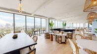 Located on an upper floor in Miami's NW 20th Street corridor, this sophisticated amenity lounge offers a generously sized, single-level space designed for both socializing and dining. Floor-to-ceiling glass windows wrap the perimeter, flooding the interior with abundant natural light and framing panoramic views of the Miami skyline and lush tree canopy below.

The open-plan layout is anchored by a striking, oversized oval bar island finished in white stone with a dark curved base, complemented by sleek black-and-gold bar stools beneath a trio of globe pendant lights. Adjacent to the bar, a dedicated dining area features a long, dark communal table surrounded by woven rattan bistro chairs, with a vibrant tropical-motif accent wall and a mounted display screen adding a bold design focal point.

The lounge area is elegantly furnished with cream upholstered armchairs and curved sofas arranged around geometric gold and black side tables, accented by tall tropical bird-of-paradise plants that reinforce the Miami aesthetic. Light wood-toned flooring flows throughout, unifying the space. Woven pendant lights and brass accents add warmth, while glass-partitioned sections offer more intimate seating zones. The overall ambiance is modern, tropical, and refined.
