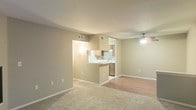 This single-story space showcases an open-concept design that connects the living area to the kitchen, creating a functional and cohesive layout. The living area features plush carpeting in a warm neutral tone, providing a comfortable foundation, while a ceiling fan overhead ensures year-round comfort. Smooth, beige-toned walls and white baseboards run throughout, contributing to a clean and consistent aesthetic.

The kitchen is partially separated from the living space by a half-wall pass-through counter, which serves both as a visual divider and a convenient surface for casual dining or food prep. Inside the kitchen, medium-toned wood cabinetry lines the walls, complemented by a stone tile backsplash that adds texture and visual interest. White appliances, including a refrigerator, are visible, and the hard flooring in the kitchen contrasts with the carpeted living area, clearly delineating the two zones. HVAC vents are positioned in the ceiling throughout, supporting efficient climate control. The overall ambiance is warm, understated, and move-in ready, with a straightforward layout that maximizes usable space.