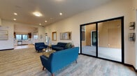 Located at 930 W 17th Ave in Junction City, Oregon, this single-story community clubhouse serves as the social hub of the Country Crossroads development. The space features a contemporary design characterized by crisp white walls, wood-tone accents, and durable plank-style flooring throughout.

The welcoming lounge area is furnished with tufted navy blue sofas and armchairs arranged around a glass-and-gold coffee table, creating an inviting gathering space. Large sliding glass doors open to an exterior courtyard, allowing natural light to flow freely into the room. Framed wall accents and decorative mirrors add warmth to the neutral palette.

A built-in display wall with warm wood shelving and white cabinetry provides attractive storage and display space, seamlessly connecting to a well-appointed kitchen. The kitchen features dark upper cabinetry, light lower cabinets, teal subway tile backsplash, white countertops, and stainless steel appliances, including a range, dishwasher, and refrigerator. A large window over the sink offers views of the surrounding area.

The game room is a standout feature, equipped with arcade machines including Pac-Man, a foosball table, and a full-size pool table. A cozy built-in window bench provides additional seating. The clubhouse also includes a property map display area near the entrance, offering residents an informative overview of the community.