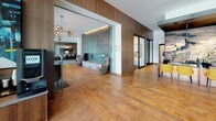 Located at 15400 NE 20th St in Bellevue, WA, this single-floor leasing and amenity lobby presents a polished, contemporary design tailored to make a strong first impression. The space is anchored by warm cork and hardwood flooring that flows throughout, complemented by rich wood-paneled accent walls and high ceilings that lend an open, airy atmosphere.

The reception area is a standout feature, boasting a dark granite-topped counter backed by a dramatic full-wall mural of Mount Rainier set against a starry night sky. Vibrant yellow upholstered barstools line the counter, adding a bold pop of color. Pendant lighting overhead enhances the striking visual effect.

Multiple distinct lounge areas are thoughtfully arranged throughout the space. One seating nook features tan leather sofas accented with deep red pillows, illuminated wall display boxes, and floor-to-ceiling windows that flood the area with natural light and views of the exterior streetscape. Another lounge area showcases rich brown leather seating, a colorful striped area rug, large-format artwork, and a sculptural multi-globe chandelier. A quieter seating alcove with a built-in fireplace offers a more intimate retreat, framed by glass partitions and warm pendant lighting. Additional private rooms with glass-panel doors suggest conferencing or meeting capabilities, rounding out this well-appointed and functional lobby environment.