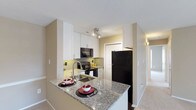 This well-appointed 3-bedroom, 2-bathroom single-floor residence offers a thoughtfully designed layout that balances comfort and functionality. The kitchen serves as a centerpiece of the home, showcasing granite countertops, a peninsula island ideal for casual dining or entertaining, and sleek white cabinetry that reaches toward the ceiling for ample storage. Black appliances, including a refrigerator, range, and built-in microwave, provide a bold contrast against the light cabinetry, while a subway tile backsplash adds a clean, modern touch. A stainless steel sink with a contemporary arc faucet anchors the workspace, and track lighting overhead ensures the area is well-lit. The kitchen flows seamlessly into the adjacent living areas, creating an open, connected feel throughout the home. Neutral-toned walls and light carpeting extend through the corridors and living spaces, lending a warm and inviting ambiance. The residence also benefits from natural light streaming in through windows visible in the adjoining rooms, further enhancing the bright and airy character of the space. With three bedrooms and two full bathrooms, this home offers generous accommodations suited for a variety of lifestyles.