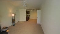 This single-floor space features an open-concept layout that seamlessly connects the living area to the kitchen. The living area is finished with neutral-toned wall-to-wall carpeting, creating a warm and understated atmosphere. A white ceiling fan is centrally positioned overhead, providing both air circulation and a subtle design element to the space.

The kitchen is partially separated by a half-wall partition, offering definition between the two areas while maintaining an open feel. It is equipped with dark wood cabinetry, a stainless steel dishwasher, and a countertop range. A track lighting system installed above the kitchen provides focused illumination across the cooking and prep areas.

Multiple doors are visible throughout the space — one near the kitchen likely leads to a pantry or utility closet, while others suggest access to additional rooms or storage. The walls are painted in a clean, neutral off-white tone throughout, contributing to a bright and cohesive interior. Overall, the space offers a functional and straightforward layout well-suited for everyday living.