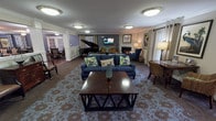 This single-floor communal living space is thoughtfully designed to foster comfort and social engagement. The layout is expansive and open, seamlessly connecting multiple functional zones beneath crown-molded ceilings with soft, ambient flush-mount lighting throughout.

At the heart of the space, a generously sized seating area anchors the room with a pair of navy blue sofas arranged around a dark wood coffee table atop a decorative floral-patterned area rug. A gas fireplace serves as a warm focal point along one wall, flanked by a wall-mounted television and framed nature-inspired artwork, including striking bird prints that lend a classic, refined character to the décor.

A polished black grand piano occupies a dedicated corner, adding an element of cultural sophistication and providing an intimate performance area. Adjacent to this, a dining area with traditional dark wood tables and upholstered chairs opens through white structural columns, offering a graceful transition between the lounge and dining spaces.

Natural light filters in through multiple windows and a set of French doors dressed with neutral grayish-beige curtains, providing views to the outside and a connection to the outdoors. Cozy reading nooks with armchairs and side tables are tucked along the perimeter, creating private retreats within the larger communal setting. The overall ambiance is warm, elegant, and inviting — well-suited for both quiet reflection and lively social gatherings.