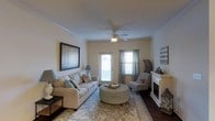 This single-floor residence offers a thoughtfully designed living space that balances comfort and style. The living room serves as the heart of the home, featuring a classic white fireplace that anchors the room with warmth and elegance. Crown molding lines the ceiling, adding a refined architectural detail throughout the space. A ceiling fan with integrated lighting provides both air circulation and ambient illumination, complementing the natural light that streams in through multiple windows dressed with layered curtains and blinds. The rich, dark hardwood flooring contrasts beautifully with the warm neutral-toned walls, creating a welcoming and timeless aesthetic. A glass-panel door provides additional brightness and offers access to the exterior. The living area is generously sized, comfortably accommodating a full sofa, accent chair, and a central ottoman, all set atop a patterned area rug that defines the gathering space. Wall art and decorative accents contribute to the overall inviting ambiance. The layout and design elements of this residence work cohesively to create a space that is both functional and visually appealing, ideal for everyday living and entertaining.
