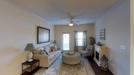 This single-floor residence offers a thoughtfully designed living space that balances comfort and style. The living room serves as the heart of the home, featuring a classic white fireplace that anchors the room with warmth and elegance. Crown molding lines the ceiling, adding a refined architectural detail throughout the space. A ceiling fan with integrated lighting provides both air circulation and ambient illumination, complementing the natural light that streams in through multiple windows dressed with layered curtains and blinds. The rich, dark hardwood flooring contrasts beautifully with the warm neutral-toned walls, creating a welcoming and timeless aesthetic. A glass-panel door provides additional brightness and offers access to the exterior. The living area is generously sized, comfortably accommodating a full sofa, accent chair, and a central ottoman, all set atop a patterned area rug that defines the gathering space. Wall art and decorative accents contribute to the overall inviting ambiance. The layout and design elements of this residence work cohesively to create a space that is both functional and visually appealing, ideal for everyday living and entertaining.