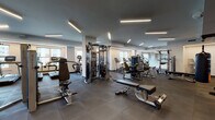 This expansive fitness center is thoughtfully designed to accommodate a wide range of workout styles within a single, open floor plan. The space is divided into distinct functional zones, each outfitted with professional-grade equipment. The main exercise floor features durable rubber tile flooring throughout, providing both traction and impact absorption, while rows of linear LED fixtures mounted to the grey ceiling deliver consistent, bright illumination across the entire area.

The cardio zone is positioned along a windowed perimeter wall, where Matrix upright bikes, treadmills, and elliptical machines benefit from natural light and views of the surrounding cityscape. Adjacent to this area, a comprehensive strength training section houses a full array of Matrix selectorized machines, cable systems, multi-functional cable towers, free weight benches, squat racks, and an organized dumbbell rack reflected in floor-to-ceiling mirrors. A heavy punching bag and suspension training equipment provide additional functional fitness options.

A separate studio room with warm hardwood floors, muted grey walls, recessed lighting, a mirrored accent wall, a mounted display screen, and ballet barre accommodates group fitness, yoga, cycling, and stretching activities, offering a quieter environment distinct from the main floor.