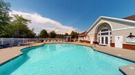 Located at 560 W New Hope Rd in Goldsboro, North Carolina, this single-story community facility serves as a central gathering space and recreational hub. The centerpiece of the property is a generously sized outdoor swimming pool with clear, blue water, surrounded by a broad concrete sundeck appointed with lounge chairs, tables, and vibrant blue patio umbrellas — ideal for relaxing on warm Carolina days. A white vinyl fence encloses the pool area, providing both safety and a clean, polished aesthetic. The clubhouse building features classic architectural details including lap siding in a neutral cream tone, a prominent arched window, and French doors that open directly onto the pool deck, creating a seamless indoor-outdoor connection. Brick accent walls at the base of the structure add a grounded, traditional character. Multiple covered patio areas are outfitted with outdoor dining tables and chairs, offering shaded spots for gatherings adjacent to the pool. Lush landscaping with mature trees and manicured shrubs borders the space, providing natural privacy and greenery. The overall ambiance is welcoming and resort-like, designed to foster community interaction and outdoor leisure in a well-maintained, attractive setting.