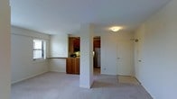 This single-floor space is designed with a functional, open layout that connects the living area to the kitchen. The living area features wall-to-wall carpeting in a neutral tone, contributing to a clean and understated aesthetic. White walls and standard-height ceilings keep the space feeling bright and straightforward.

The kitchen is positioned adjacent to the living area and is defined by warm, medium-toned wood cabinetry on both upper and lower sections. A pass-through counter with a granite-style surface serves as a visual and functional divider between the kitchen and living space, offering convenience for everyday use. The kitchen includes built-in appliances such as a microwave and a refrigerator, which is visible through a connecting doorway.

A structural column near the center of the space adds an architectural element while delineating the transition between zones. A doorway leading off the main area provides access to additional rooms. Natural light enters through a window fitted with horizontal blinds on the living area wall, supplemented by ceiling-mounted light fixtures throughout. The overall ambiance is clean, neutral, and practical.