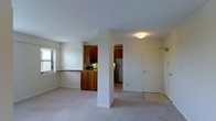 This single-floor space is designed with a functional, open layout that connects the living area to the kitchen. The living area features wall-to-wall carpeting in a neutral tone, contributing to a clean and understated aesthetic. White walls and standard-height ceilings keep the space feeling bright and straightforward.

The kitchen is positioned adjacent to the living area and is defined by warm, medium-toned wood cabinetry on both upper and lower sections. A pass-through counter with a granite-style surface serves as a visual and functional divider between the kitchen and living space, offering convenience for everyday use. The kitchen includes built-in appliances such as a microwave and a refrigerator, which is visible through a connecting doorway.

A structural column near the center of the space adds an architectural element while delineating the transition between zones. A doorway leading off the main area provides access to additional rooms. Natural light enters through a window fitted with horizontal blinds on the living area wall, supplemented by ceiling-mounted light fixtures throughout. The overall ambiance is clean, neutral, and practical.