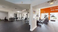 This single-floor fitness center is thoughtfully designed to accommodate a wide range of workout styles across several distinct zones. The main cardio area makes a strong visual impact with its bold orange accent wall rising to impressive heights, adorned with motivational phrases such as "Whatever You Do, Do Not Give Up!" Twin flat-screen TVs are mounted high on the wall, and large floor-to-ceiling windows flood the space with natural light while offering views of the surrounding greenery. The rubber-floored cardio zone is lined with ellipticals, treadmills, and a rowing machine. Adjacent to this, the strength training area features a comprehensive free weight section with a full dumbbell rack, adjustable benches, and wall-mounted mirrors for form checks. A dedicated functional fitness zone includes a turf section with a punching bag, medicine balls, battle ropes, and plyo boxes, accompanied by motivational wall lettering. A separate, quieter room with light wood-style flooring serves as a flexibility and mind-body space, complete with stability balls, a BOSU trainer, and a wall-mounted TV. Throughout the facility, durable rubber flooring, textured ceilings, and consistent white walls with energizing orange accents create a cohesive, motivating atmosphere.